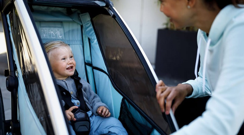 Kind sitzt sicher angeschnallt im Fahrrad-Kinderanhänger, während eine Erwachsene den Anhänger vorbereitet – ideal für komfortable Familienausflüge mit dem Fahrrad