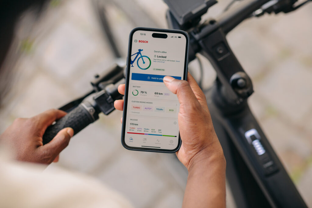 Smartphone mit Bosch eBike Flow App in der Hand über einem E-Bike mit integriertem Bosch Kiox 400C Display, zeigt Akkustand, Reichweite und Fahrmodi für vernetztes E-Bike-Management
