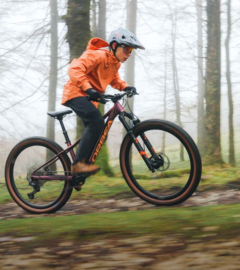 Das Orbea Kimu 27 Kinderfahrrad. Das Kindermountainbike für Kinder im Alter von 9 bis 12 Jahren.
Ein echtes Mountainbike für Kinder. Mit 110mm Federweg in der Federgabel einer Teleskopsattelstütze und großen 27,5 Zoll Reifen.
Im Bild fährt ein Junge mit oranger Jacke das neue Orbea Kimu 27. Er fährt im Wald und trägt einen grauen Helm.