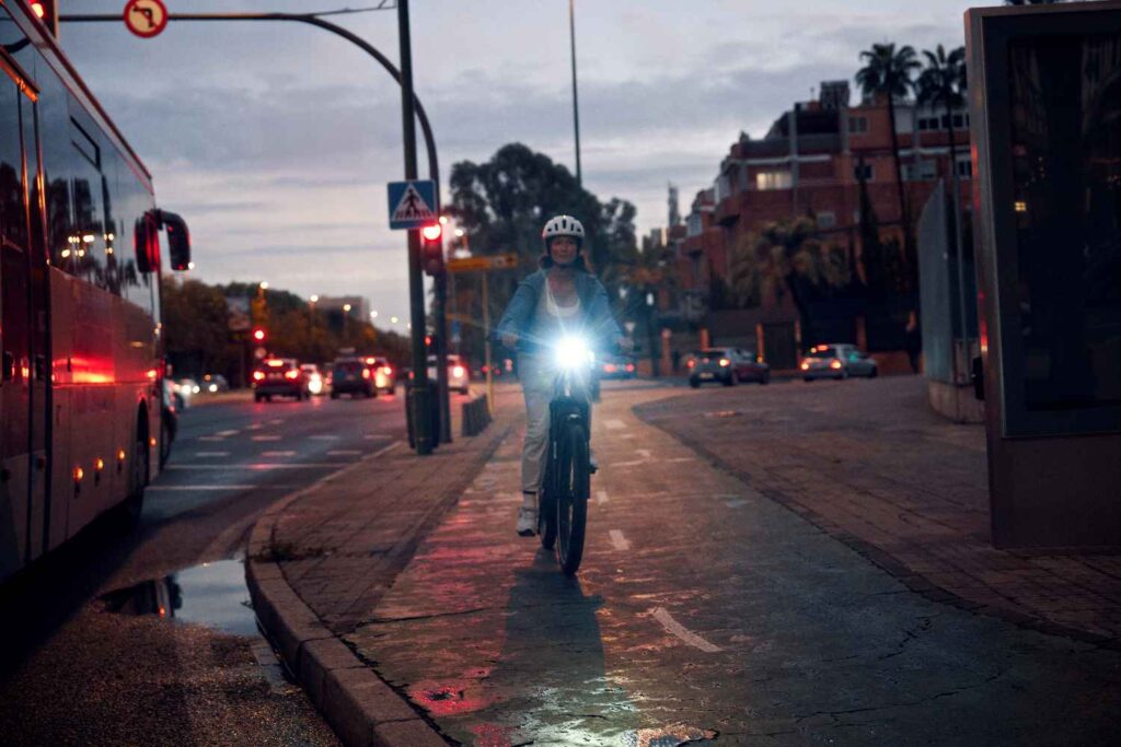 Fahrradausstattung für Pendler.
Auf dem Bild ist eine Frau auf einem E-Bike zu erkennen die mit einem eingeschalteten Fahrradlicht in der Stadt fährt