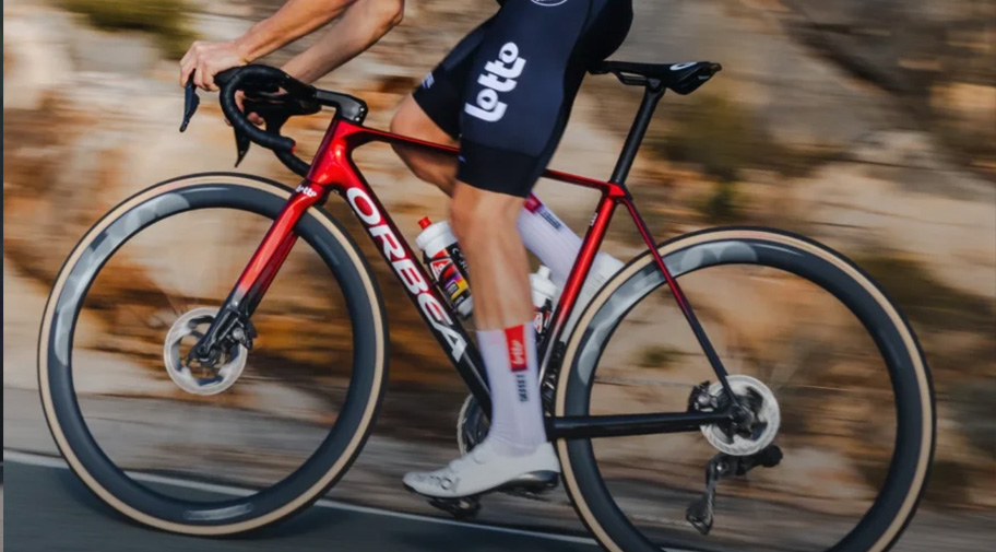 Ein Straßenradfahrer in rotem Trikot und weißem Helm ist auf einem modernen Rennrad mit tiefen Carbon-Laufrädern unterwegs. Durch die Bewegungsunschärfe im felsigen Hintergrund wird die Dynamik und das hohe Tempo der Fahrt hervorgehoben