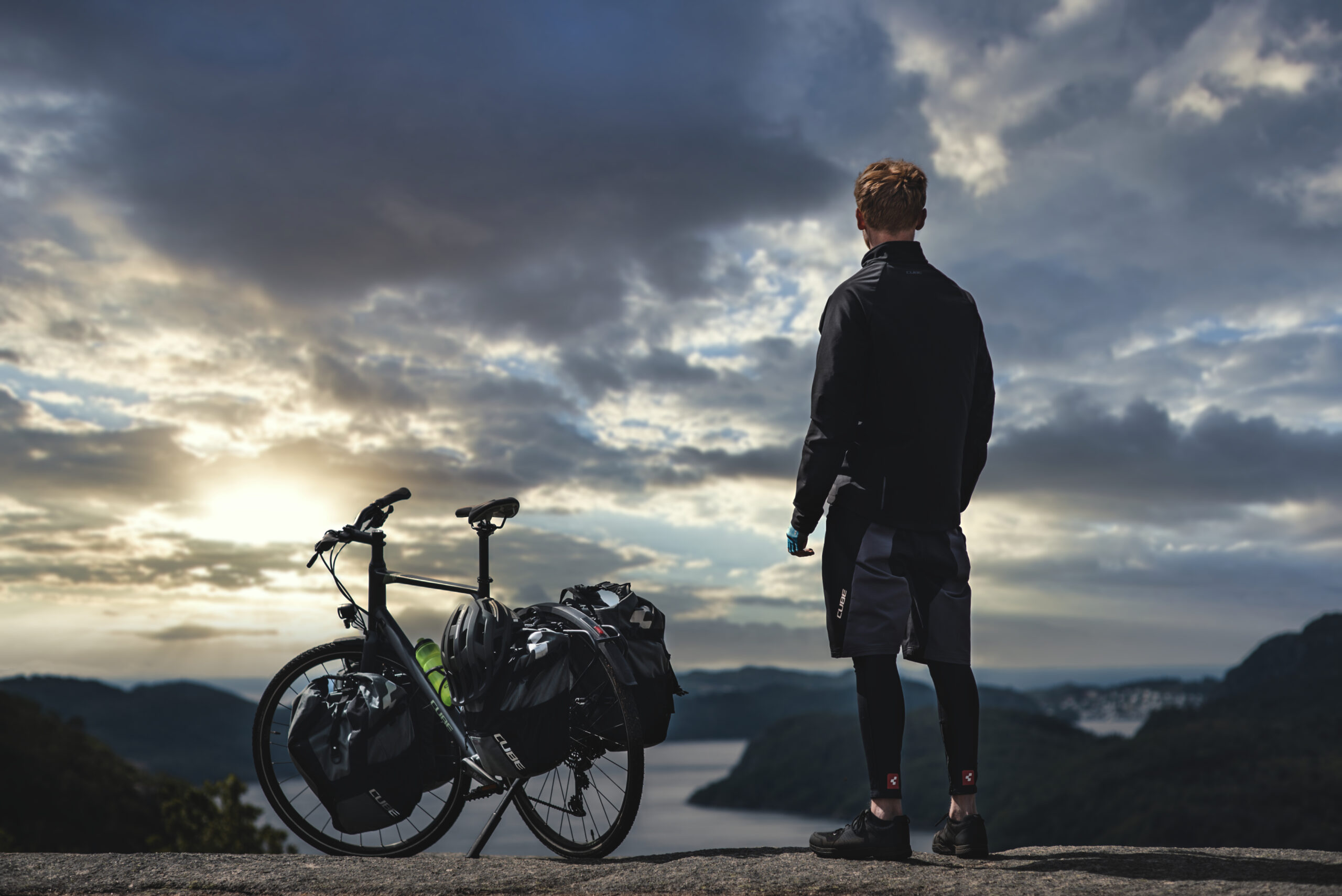 Radreisender steht mit seinem bepackten Cube Trekkingrad auf einer Anhöhe und blickt auf eine Landschaft bei Abendlicht