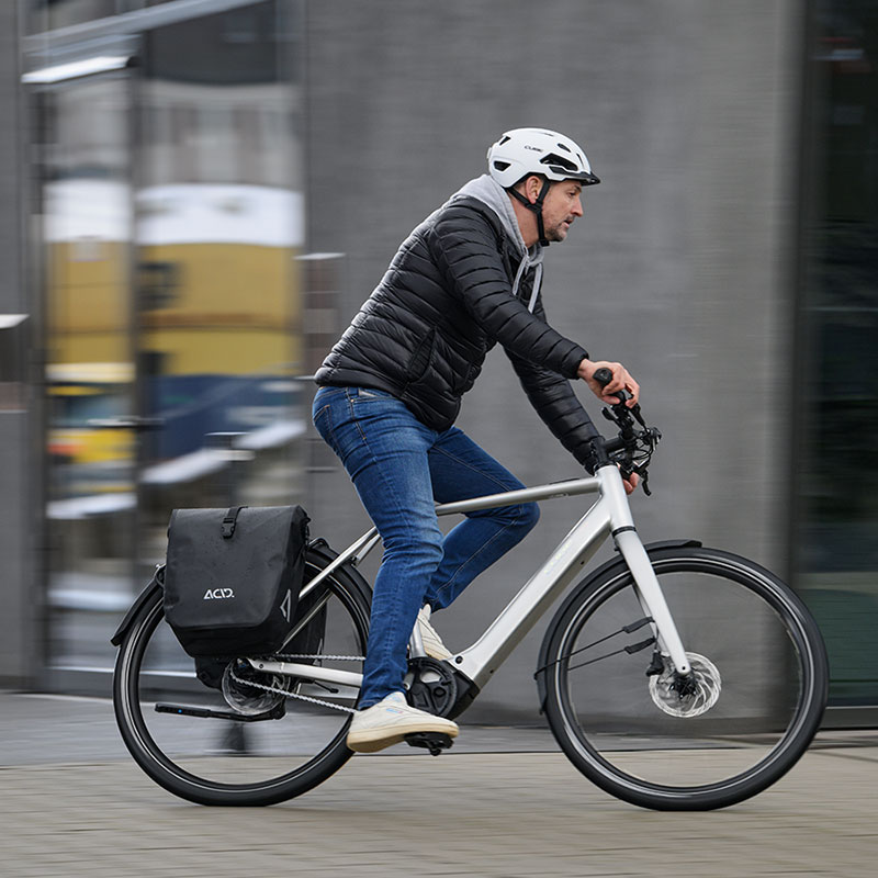 Eine Person fährt mit einem silbernen E-Bike auf einer gepflasterten Straße in einer städtischen Umgebung. Sie trägt einen weißen Fahrradhelm, eine schwarze Steppjacke, Jeans und transportiert eine schwarze Gepäcktasche am Hinterrad.
