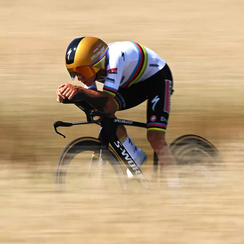 Ein Radrennfahrer in Weltmeistertrikot fährt auf einem Zeitfahrrad in aerodynamischer Haltung mit hoher Geschwindigkeit über eine Straße. Der unscharfe Vorder- und Hintergrund betont die Dynamik der Szene.