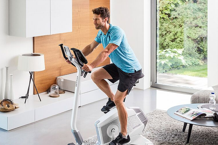 Mann mit blauem Oberteil, der in einem Wohnzimmer auf einem Ergometer trainiert