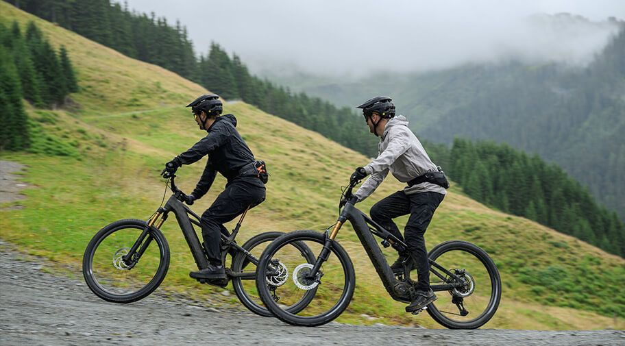 Dieses Foto zeigt zwei Radfahrer, die auf elektrischen Mountainbikes eine bergige, nebelverhangene Landschaft entlangfahren.