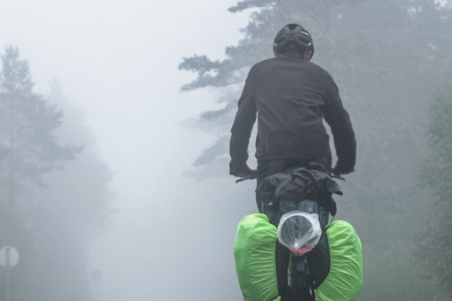 Radfahrer in dunkler Kleidung und mit reflektierendem Gepäck, der durch dichten Nebel fährt.